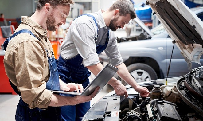 Side view portrait of two modern bearded mechanics looking under hood of car inspecting engine and using laptop  while working in car service and repair center, copy space